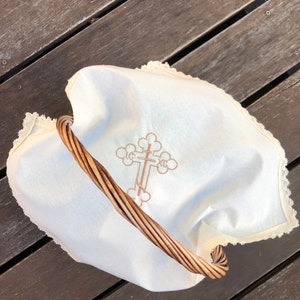 May include: A white fabric basket liner with a brown wicker handle and an embroidered cross design. The liner has a lace trim around the edges.