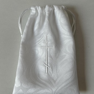 May include: White drawstring bag with a white embroidered cross. The bag has a white floral pattern.