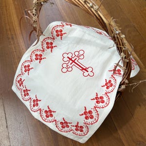 Op de afbeelding: Een witte doek met een rood geborduurd kruis en bloemenrand, geplaatst in een gevlochten mand met een handvat. De mand is gemaakt van bruine twijgen en versierd met kleine witte kralen en gedroogde bloemen.