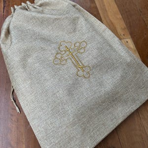 May include: A beige burlap drawstring bag with a gold embroidered cross design. The bag is made of a textured fabric and is closed with a drawstring. The cross is centered on the front of the bag.