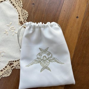 Puede incluir: Una bolsa blanca con cordón y un diseño de ángel bordado en oro. La bolsa tiene un diseño simple y elegante, con un cierre de cordón dorado. Un tapete de encaje blanco es parcialmente visible en el fondo.