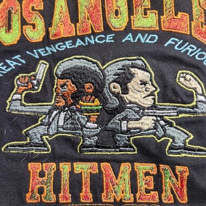 May include: Black t-shirt with a red, green, and yellow embroidered design. The design features two cartoon figures holding guns and the text "Los Angeles", "Great Vengeance and Fury", "Hitmen", and "Estd 1994".