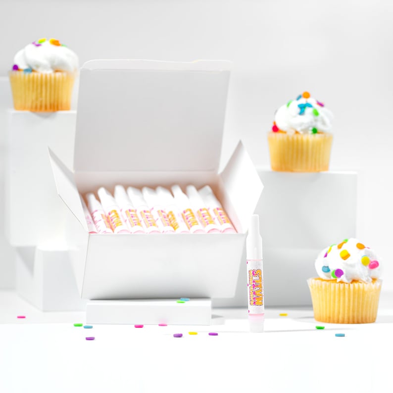 May include: A white box containing a selection of colourful frosting tubes. The box is open, revealing the tubes. There are also three cupcakes topped with white frosting and colourful sprinkles. The cupcakes are displayed on white surfaces.