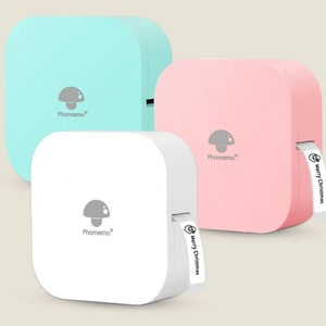May include: Three portable label makers in different colours: light blue, pink and white. Each label maker has a mushroom logo and the text "Phomemo" printed on the front. The pink label maker has a white label with the text "Merry Christmas" printed on it.