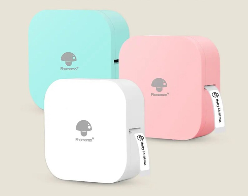 May include: Three portable label makers in different colours: light blue, pink and white. Each label maker has a mushroom logo and the text "Phomemo" printed on the front. The pink label maker has a white label with the text "Merry Christmas" printed on it.