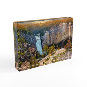 May include: A rectangular acrylic block featuring a scenic view of Yellowstone National Park. The image showcases a waterfall cascading into a canyon, surrounded by trees and cliffs. The text "YELLOWSTONE NATIONAL PARK" is visible.