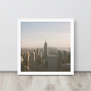 Puede incluir: Una impresión enmarcada que presenta una vista aérea del horizonte de la ciudad de Nueva York. El Empire State Building se muestra de forma destacada, rodeado de otros rascacielos. La imagen tiene una paleta de colores suaves, en un marco blanco.