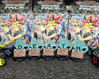 Op de afbeelding: Een verzameling Yu-Gi-Oh! booster packs en decoratieve items. De items bevatten turquoise gekleurde, gestileerde letters die "DEATH" spellen, kleine bronskleurige plaques en zwarte dobbelstenen met kleurrijke symbolen.