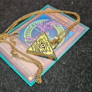 May include: A gold chain necklace with a gold pendant shaped like the Eye of Horus, a symbol from Egyptian mythology. The pendant is resting on a blue and white card with the text "MAGICIAN" and "ATK/2500 DEF/2100".