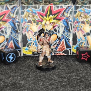 Puede incluir: Una figura en miniatura de un guerrero frente a tres cajas de cartas de Yu-Gi-Oh!. Las cajas presentan personajes de anime con colores vibrantes. Dados con símbolos a ambos lados de la figura. La figura está pintada con detalles.