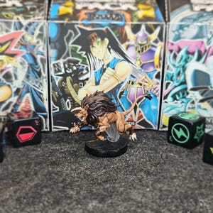 May include: A miniature brown lion figure with a dark mane stands on a black circular base, positioned in front of three Yu-Gi-Oh! booster pack boxes. Four black six-sided dice with coloured symbols, including blue swords, a red hexagon, a green lightning bolt, and a yellow arrow, are arranged around the central lion figure.