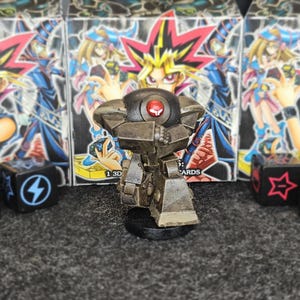 Puede incluir: Una detallada figura de aspecto metálico de un robot con un ojo rojo, de pie sobre una base negra. El fondo presenta múltiples cajas de cartas coleccionables de Yu-Gi-Oh! y varios dados con símbolos.