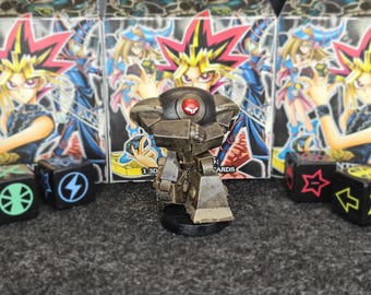 Puede incluir: Una detallada figura de aspecto metálico de un robot con un ojo rojo, de pie sobre una base negra. El fondo presenta múltiples cajas de cartas coleccionables de Yu-Gi-Oh! y varios dados con símbolos.