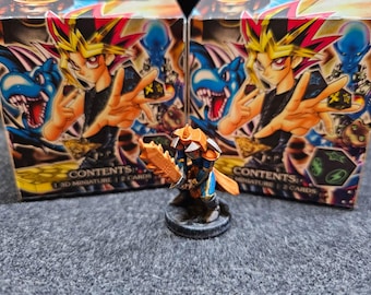 Puede incluir: Dos cajas sin abrir de cartas de intercambio de Yu-Gi-Oh! con un monstruo azul y amarillo en la parte delantera. Las cajas tienen el texto "Contenido: 1 miniatura 3D | 2 cartas" en la parte inferior. Una miniatura pintada de un guerrero azul y dorado con una espada está en primer plano.