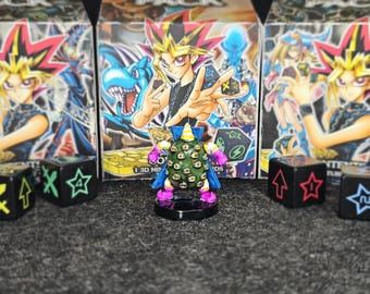 Peut inclure: Une figurine miniature colorée avec de nombreux yeux, sur une base noire. Des dés noirs avec des symboles colorés et des boîtes de jeu de cartes sont en arrière-plan. Le thème est un jeu de cartes.