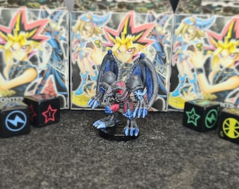 Puede incluir: Una figura en miniatura de una criatura alada y con cuernos con armadura de color gris oscuro, azul y rojo, sobre una base circular negra. La figura presenta garras afiladas y alas grandes, colocada frente a tres cajas temáticas de Yu-Gi-Oh! y cuatro dados negros.