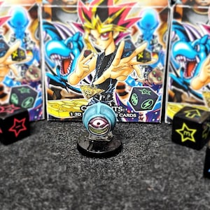 May include: A blue, one-eyed monster figurine on a black base, with black dice featuring symbols and card packs with a character with spiky yellow hair. The dice have star, lightning, and other symbols.