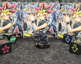 Peut inclure: Une figurine de dragon noir avec des détails rouges, posée sur une base noire. Le fond présente plusieurs boîtes avec des illustrations de style anime et des dés avec des symboles. Un objet de collection.