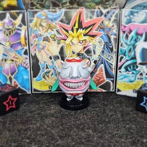 Peut inclure: Une figurine Yu-Gi-Oh! Pot de Cupidité représentant un visage rond et blanc souriant avec des dents proéminentes et des accents rouges, placée devant une boîte de cartes Yami Yugi. Deux dés noirs avec des étoiles et flèches colorées.