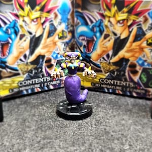 May include: A purple and yellow painted miniature figurine of a jester with a long, curled tail. The figurine is standing on a black base with a textured surface. The figurine is a 3D miniature.