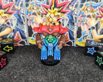 May include: A hand-painted miniature figure of a creature with a red shoulder armor piece, brown arms, and a blue lower body section. The figure features a small green character seated in the center, with a gold-colored arch detail on top.