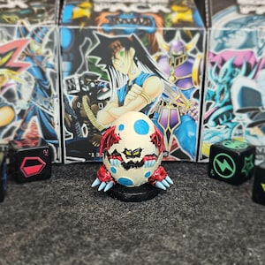 Può includere: Una piccola statuetta a forma di uovo color crema con macchie blu, ali rosse e artigli affilati, posizionata su una base circolare nera. Quattro dadi neri con simboli colorati circondano la figura, davanti a scatole a tema Yu-Gi-Oh!.