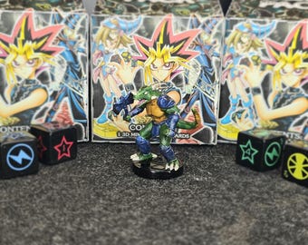Puede incluir: Una pequeña figura de criatura reptiliana verde con armadura azul y un hacha oscura, de pie sobre una base circular negra. En el fondo, tres cajas de producto idénticas muestran un personaje de anime con cabello amarillo puntiagudo, y cuatro dados negros con símbolos de colores están colocados alrededor de la figura.