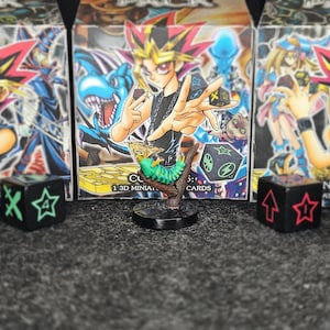 May include: A 3D miniature of a green caterpillar on a branch, with a character from a card game in the background. Several black dice with white, red, and blue symbols are in front of the miniature. Card game boxes are behind the dice.