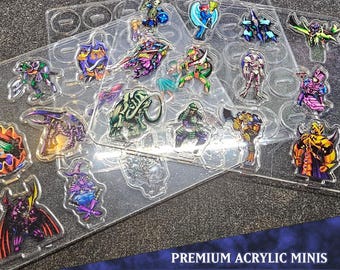 May include: Clear acrylic mini figures of various monsters in a Monster Pack 5 set. The figures are colorful, with designs including a samurai, a wizard, and other fantasy creatures. The text "PREMIUM ACRYLIC MINIS" and "MONSTER PACK 5" are visible.
