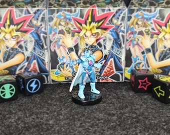 Puede incluir: Una figura de acción azul claro se encuentra sobre una base negra, con una capa blanca y detalles plateados. La figura está frente a tres cajas con ilustraciones de estilo anime y dados con símbolos. Las cajas dicen "CARDS".