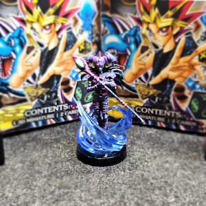 Könnte beinhalten: Eine bemalte Miniaturfigur eines dunkelvioletten und blauen Charakters, der einen Stab mit einer blauen und grünen Kugel oben hält. Der Charakter steht auf einem blauen und weißen Sockel mit einem wirbelnden blauen Effekt. Die Miniatur ist von zwei Schachteln mit dem Text "CONTENTS: 1 3D Miniature | 2 Cards" umgeben.