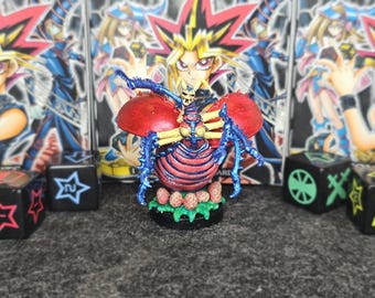 May include: A detailed figurine of a Yu-Gi-Oh! monster, featuring a red body with blue and yellow accents. The figure has a skull-like head and is set against a backdrop of Yu-Gi-Oh! character cards and dice.