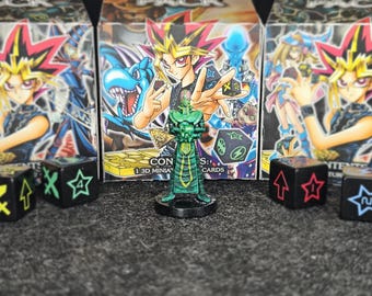 May include: A green miniature figure of a character from the Yu-Gi-Oh! trading card game, standing in front of card packs. Black dice with white symbols are in the foreground. The card packs feature anime-style illustrations.