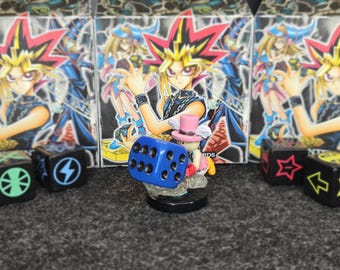 Puede incluir: Un dado azul con puntos blancos y una pequeña figura con sombrero rosa y alas están frente a tres cajas de cartas Yu-Gi-Oh! Las cajas presentan personajes de anime. Dados negros con símbolos a cada lado.