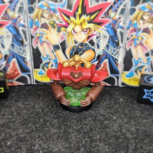 Op de afbeelding: Een rood, bruin en goudkleurig beeldje van een personage uit de Yu-Gi-Oh! anime-serie, met een groene bol. De achtergrond bevat meerdere Yu-Gi-Oh! karakterkaarten en zwarte dobbelstenen met witte symbolen.