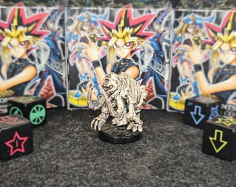 May include: A detailed, off-white skeletal monster figurine stands on a black base. The figurine is in front of several Yu-Gi-Oh! card boxes and black dice with colorful symbols. The background is a dark gray surface.