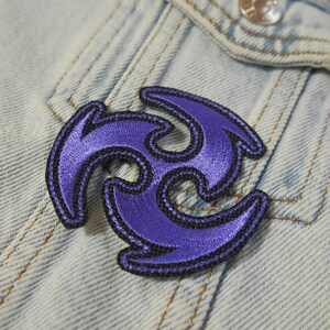 Puede incluir: Parche bordado en color morado y negro con forma de estrella de tres puntas. El parche est&aacute; sobre un fondo de mezclilla azul.
