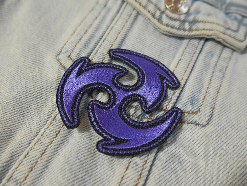 Puede incluir: Parche bordado en color morado y negro con forma de estrella de tres puntas. El parche est&aacute; sobre un fondo de mezclilla azul.