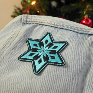 Puede incluir: Un parche en forma de copo de nieve azul claro con contornos negros cosido en una chaqueta vaquera azul claro.