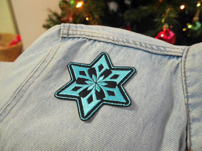 Puede incluir: Un parche en forma de copo de nieve azul claro con contornos negros cosido en una chaqueta vaquera azul claro.