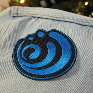 Puede incluir: Un parche bordado azul y negro con un dise&ntilde;o circular. El dise&ntilde;o presenta una serie de curvas y c&iacute;rculos superpuestos.