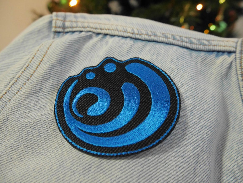 Puede incluir: Un parche bordado azul y negro con un dise&ntilde;o circular. El dise&ntilde;o presenta una serie de curvas y c&iacute;rculos superpuestos.