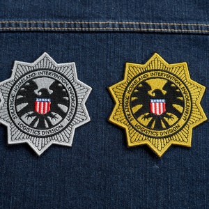 May include: Two embroidered patches with a starburst design. The patches are silver and gold with a black eagle and a red, white and blue shield. The text on the patches reads "Strategic Homeland Intervention Enforcement Logistics Division".