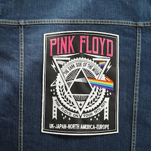 May include: A black and white embroidered patch with the text "Pink Floyd" in pink lettering. The patch features a graphic of a prism with a rainbow beam of light and the text "The Dark Side of the Moon". The patch also includes the text "Pink Floyd on Tour 1972-1973" and "UK-Japan-North America-Europe".