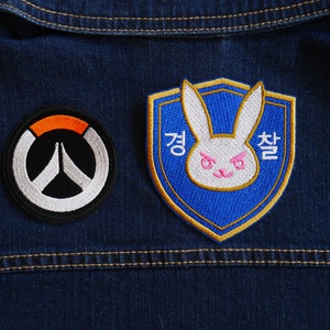 Könnte beinhalten: Zwei gestickte Aufnäher auf einer blauen Jeansjacke. Der linke Aufnäher ist ein weißes und schwarzes Overwatch-Logo mit orangefarbenem Rand. Der rechte Aufnäher ist ein blauer Schild mit einem weißen Kaninchen mit rosa Augen und Nase. Der koreanische Text "경찰" ist auf dem Schild gestickt.