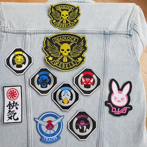 Op de afbeelding: Een spijkerjack met verschillende geborduurde patches met personages en logo's uit de videogame Overwatch. De patches bevatten Tracer, D.Va, Reaper, Genji, Soldier: 76 en Deadlock Rebels.