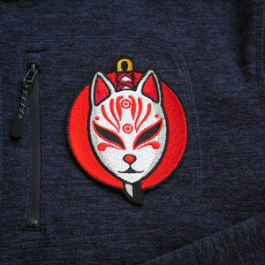 May include: Embroidered patch of a Kitsune mask in white and red, set on a red circle. The patch is sewn onto a navy blue fleece jacket, with a black zip and pocket. The jacket is a dark blue colour.