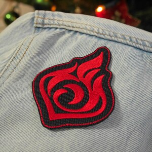 Puede incluir: Parche bordado rojo y negro con un dise&ntilde;o de llama estilizado. El parche est&aacute; cosido en una chaqueta vaquera azul claro.