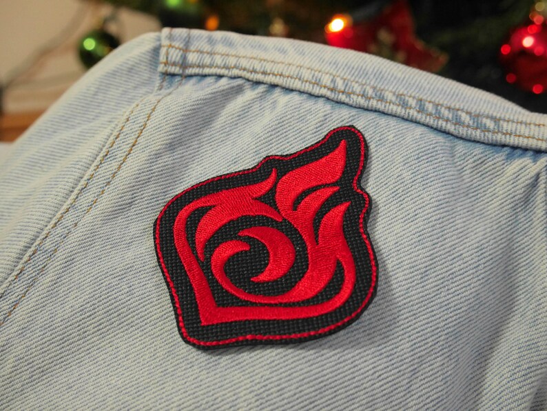 Puede incluir: Parche bordado rojo y negro con un dise&ntilde;o de llama estilizado. El parche est&aacute; cosido en una chaqueta vaquera azul claro.