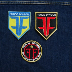 Op de afbeelding: Drie geborduurde patches met de tekst "FRINGE DIVISION" en een gestileerde "F" in verschillende kleuren. De bovenste twee patches zijn schildvormig met een gele en blauwe "F" op een blauwe achtergrond en een rode "F" op een zwarte achtergrond. De onderste patch is rond met een zwarte "F" op een rode achtergrond en de tekst "DEPARTMENT OF DEFENCE" eromheen.